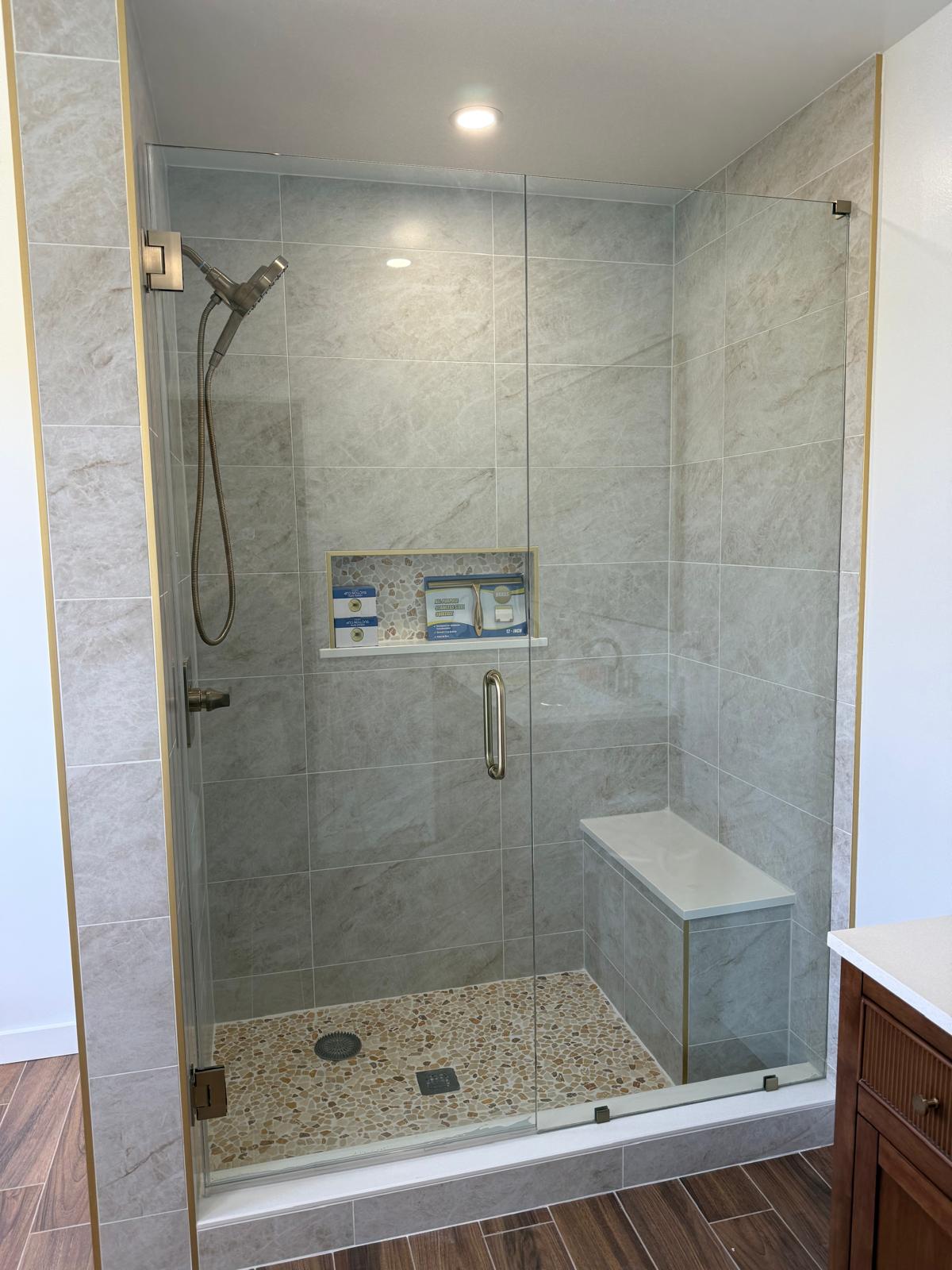 Frameless glass shower door beside gray tile and a built-in bench